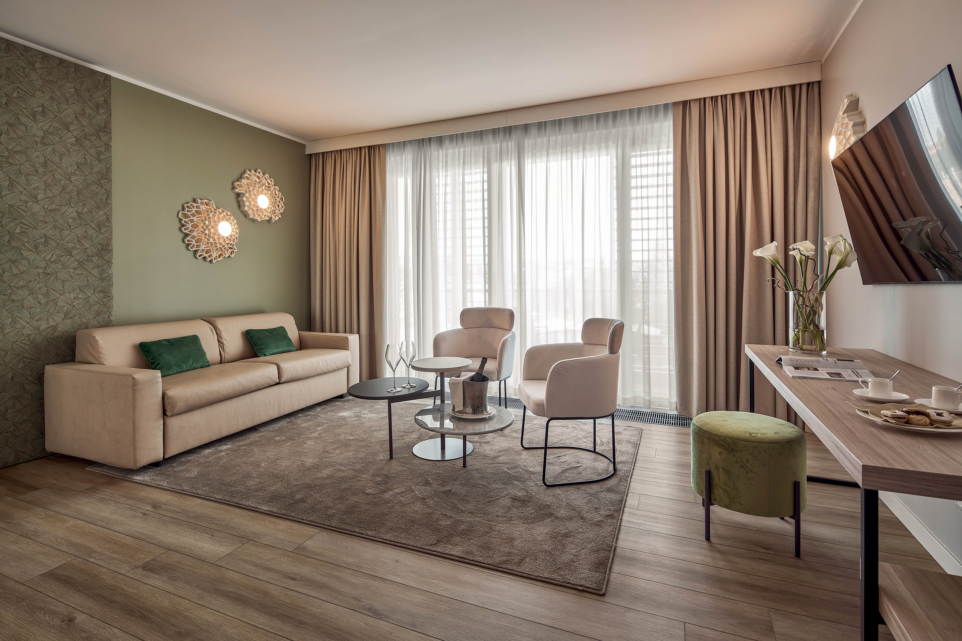 A contemporary living room features a beige sofa with green accent cushions and two light armchairs around glass coffee tables. Floor-to-ceiling windows with sheer curtains and heavier drapes fill the space with soft natural light. A wall-mounted television faces a slim console table holding flowers and tableware, while warm wall sconces add ambient lighting. The room has wood-look flooring, a large area rug, and a calm, neutral color palette with subtle green tones.
