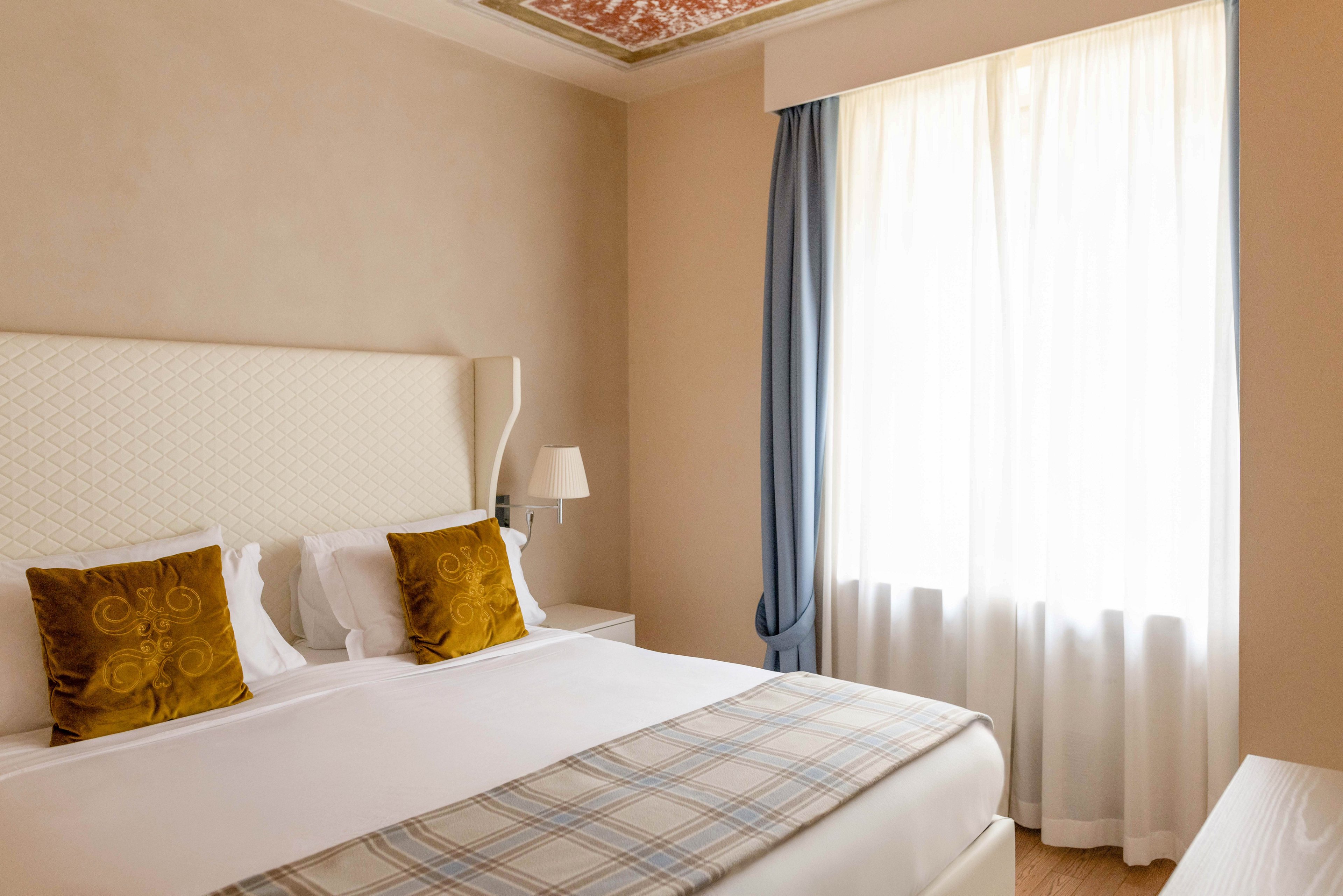 A neatly made double bed with white linens and patterned pillows stands against a light-colored wall in a cozy bedroom. Natural light streams through a large window covered with sheer curtains and framed by blue drapes. A bedside table with a lamp sits next to the bed, adding a warm and inviting touch. The overall decor is soft and neutral, creating a calm and comfortable atmosphere.