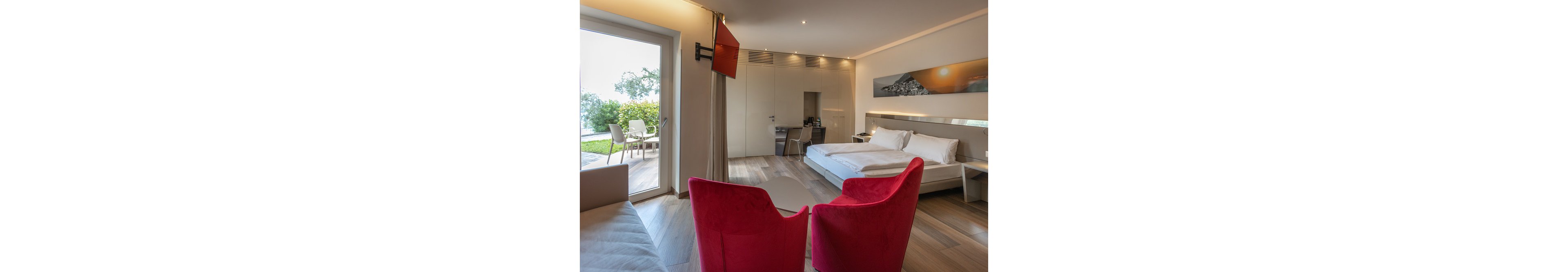 A contemporary hotel room features a large bed, two vibrant red chairs, and a wall-mounted television. The space is illuminated by natural light from a glass door leading to an outdoor patio with seating and greenery. Neutral tones dominate the palette, complemented by the bold accent chairs and a landscape artwork above the bed. The room exudes a calm, inviting atmosphere with minimalist decor and clean lines.