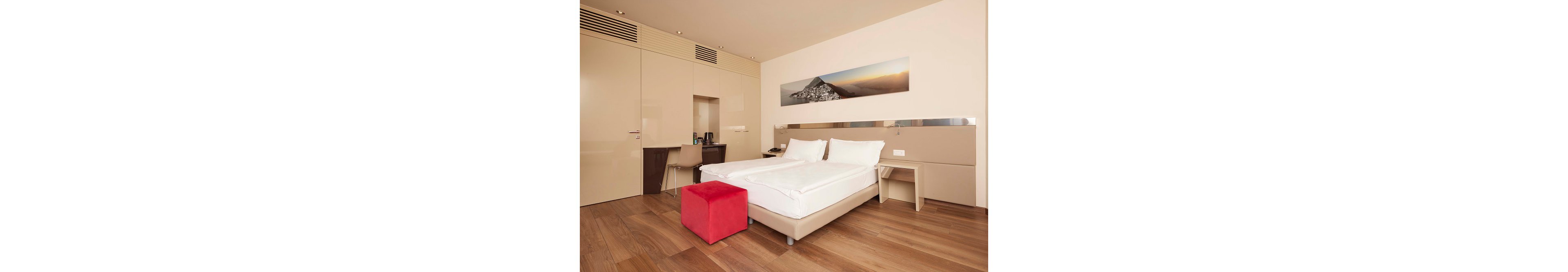 A contemporary hotel room features a double bed with crisp white linens, a red ottoman, and light wood flooring. The space includes a small desk area with a chair and coffee amenities, set against beige walls and cabinetry. Above the bed, a panoramic mountain landscape artwork adds visual interest, while the overall palette is neutral and warm.