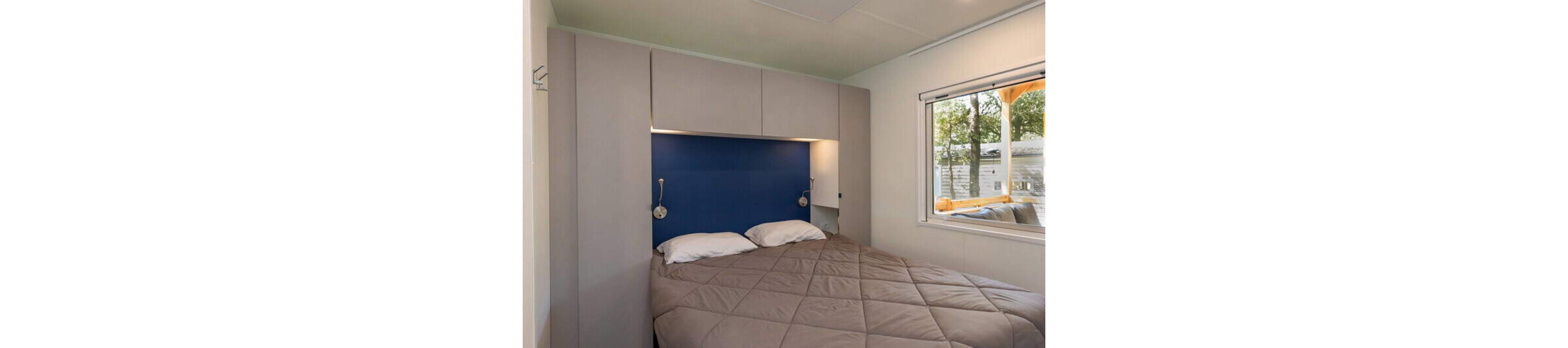 A neatly made double bed sits against a blue accent wall in a compact modern bedroom. Built-in cabinets surround the headboard, creating a streamlined and functional design. Two wall-mounted reading lights flank the bed, adding focused illumination. A large window on the right side lets in natural light and offers a view of trees and an outdoor structure.