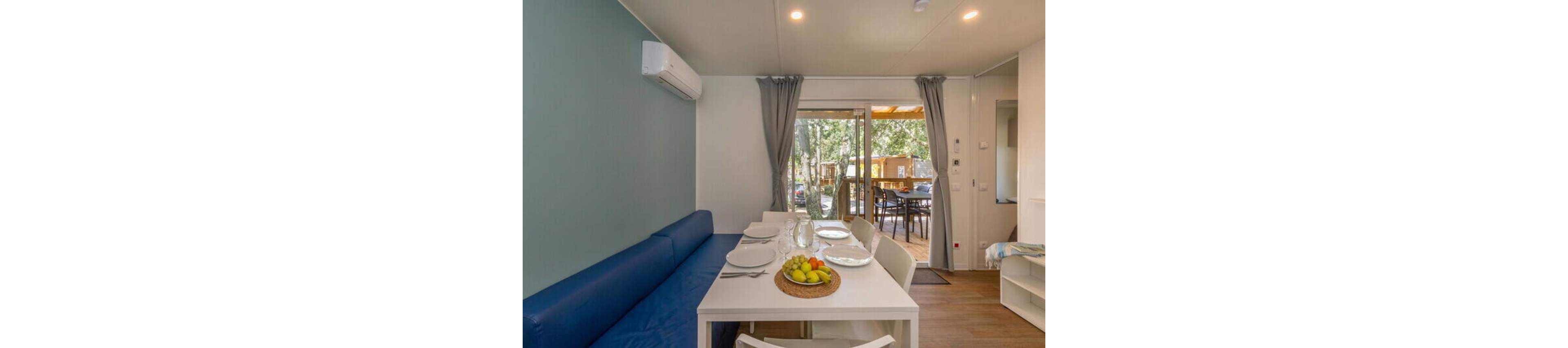 Bright interior of a modern holiday cabin features a white dining table set with plates, glasses, and a fruit centerpiece. A blue upholstered bench runs along the wall beneath an air conditioning unit, adding seating to the compact living space. Large glass doors with gray curtains open to a wooden deck with outdoor table and chairs, overlooking trees and neighboring cabins. The room has light wood flooring and simple contemporary decor, creating a clean and inviting atmosphere.