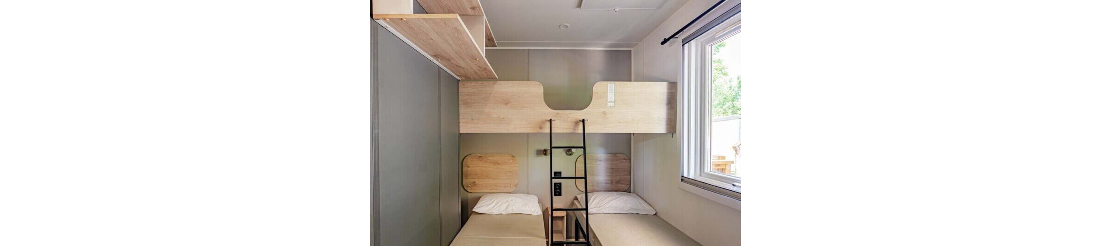 The image shows a compact bedroom with a bunk bed system featuring two lower single beds and an upper bunk accessed by a black metal ladder. Light wood panels and shelves contrast with neutral beige bedding and gray walls, creating a clean, minimalist look. A large window on the right lets in natural light and offers a view of greenery outside. The overall design suggests a small-space solution suitable for hostels, cabins, or tiny homes.