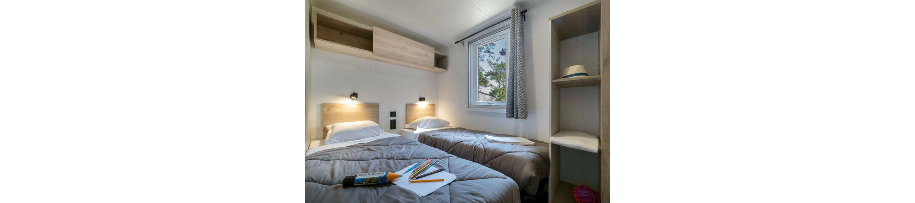 A compact bedroom features two single beds with gray comforters and wooden headboards. A small central desk holds colored pencils, a notebook, and a pencil case, suggesting a study or creative activity. Wall-mounted reading lights illuminate the beds, and a window with a patterned curtain brings in natural light. Open shelving with a hat and other items adds storage and a casual, lived-in feel to the room.