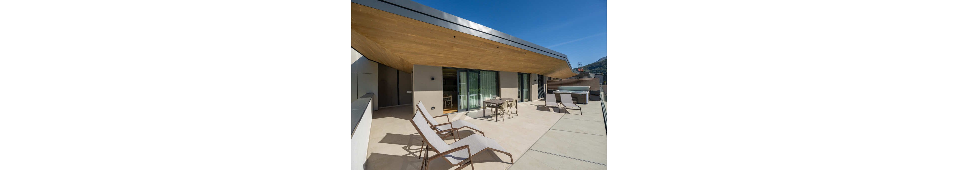 A spacious modern rooftop terrace features several reclining lounge chairs and a small dining table with chairs. Large sliding glass doors connect the terrace to the interior living space. The architecture showcases clean lines, a wood-paneled overhang, and neutral-toned flooring. Mountains and blue sky are visible in the background, emphasizing an open, outdoor lifestyle.