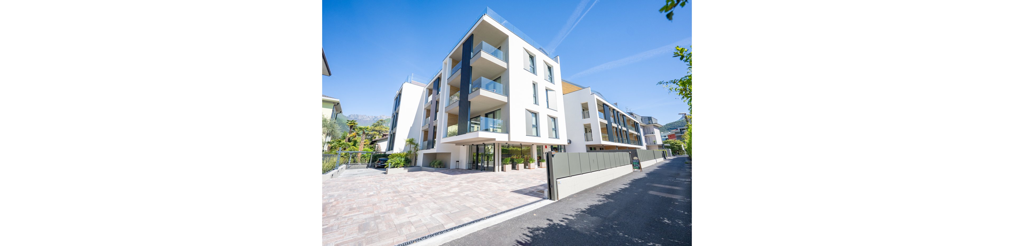 Contemporary multi-story apartment building with clean white facades and large glass balconies stands along a quiet street. The structure features geometric lines, expansive windows, and recessed terraces. A wide paved driveway and gated entrance lead to the main entry at ground level. Bright daylight and clear blue sky emphasize the modern, upscale residential setting.