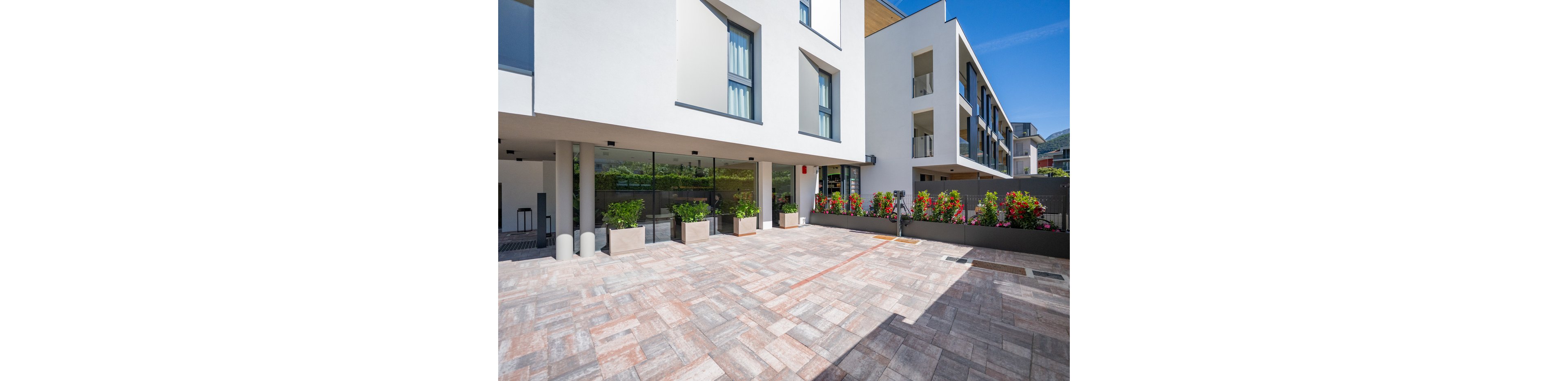 Contemporary white apartment buildings frame a spacious paved courtyard under a clear blue sky. Large glass windows and balconies overlook the open area, creating a clean and minimalist architectural look. Rectangular planters with green shrubs and red flowers line the edge of the courtyard, adding color and softness to the hardscape. The bright daylight emphasizes the geometric lines and modern urban residential setting.