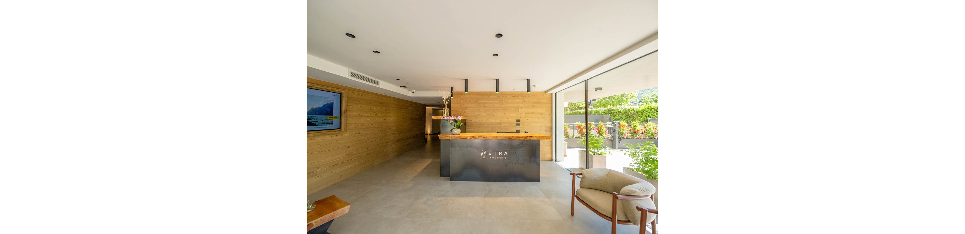 Contemporary reception area featuring a sleek front desk with the word 'NETRA' visible on its surface. The space has light-colored flooring, wooden wall paneling, and large windows that open to a green outdoor area. A comfortable armchair and a wall-mounted television add to the welcoming atmosphere. Bright natural light and minimalist decor create a clean, modern aesthetic.