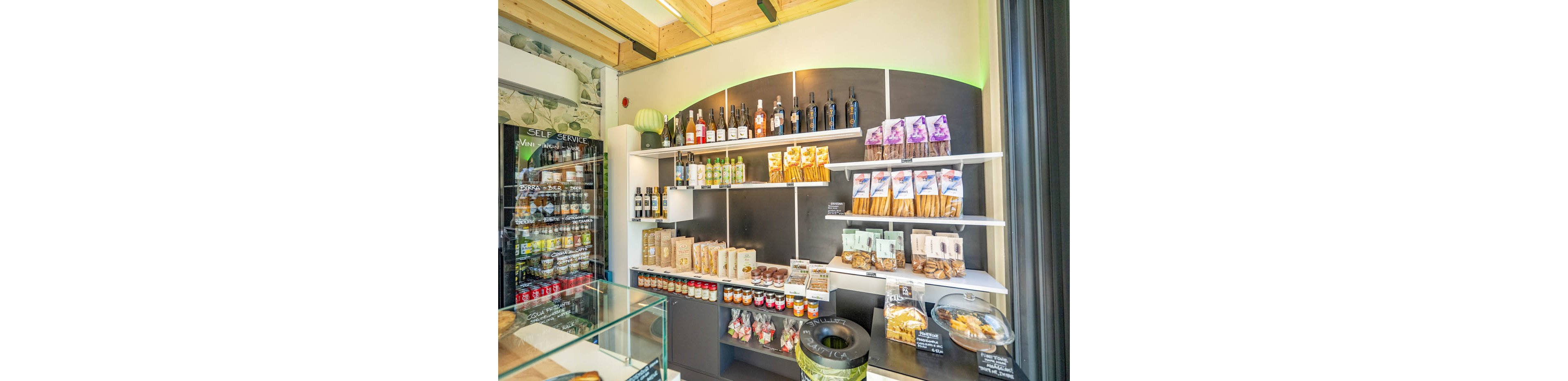 A small gourmet shop interior displays neatly arranged food and drink products on wall shelves. Bottles of wine and spirits stand on the upper shelves, while bags of snacks, breadsticks, and packaged cookies fill the middle and lower levels. A refrigerated display case and a glass counter with pastries are visible in the foreground. Bright lighting and light wood ceiling beams create a clean, contemporary atmosphere.
