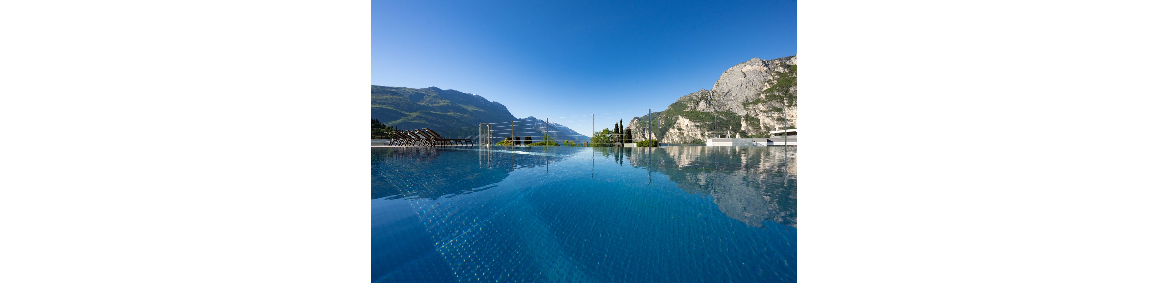 A tranquil infinity pool stretches toward a dramatic mountain range under a clear blue sky. The water surface reflects the surrounding peaks and greenery, creating a mirror-like effect. Lounge chairs line the deck on one side, suggesting a relaxing resort or spa setting. The overall scene conveys calm, luxury, and peaceful outdoor leisure.