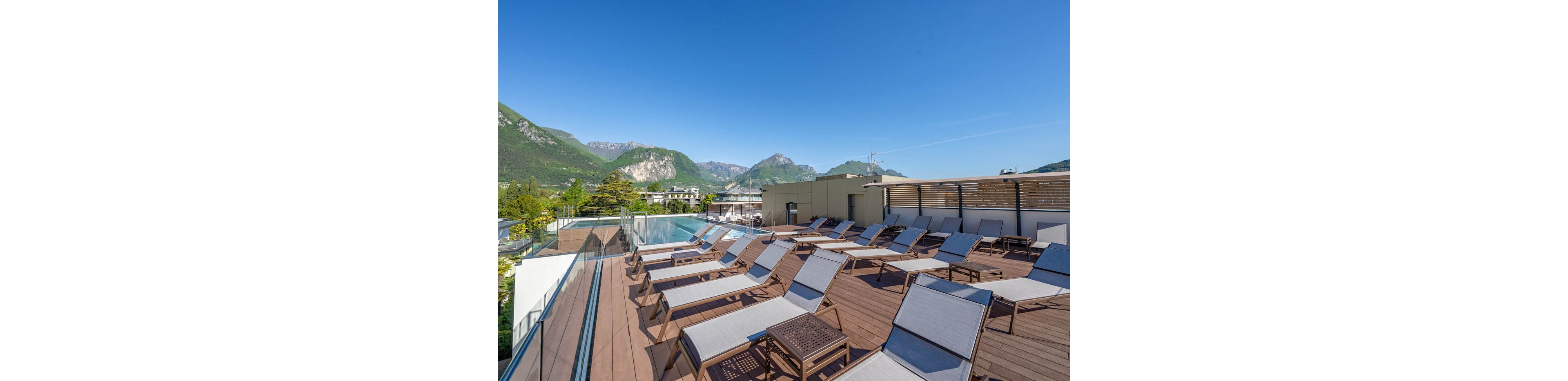 A modern rooftop terrace features numerous sun loungers arranged along a wooden deck. An infinity-style swimming pool runs beside a glass railing overlooking the surrounding area. In the background, green mountains and rocky peaks rise under a clear blue sky. The scene suggests a tranquil resort or hotel setting ideal for relaxation and sunbathing.