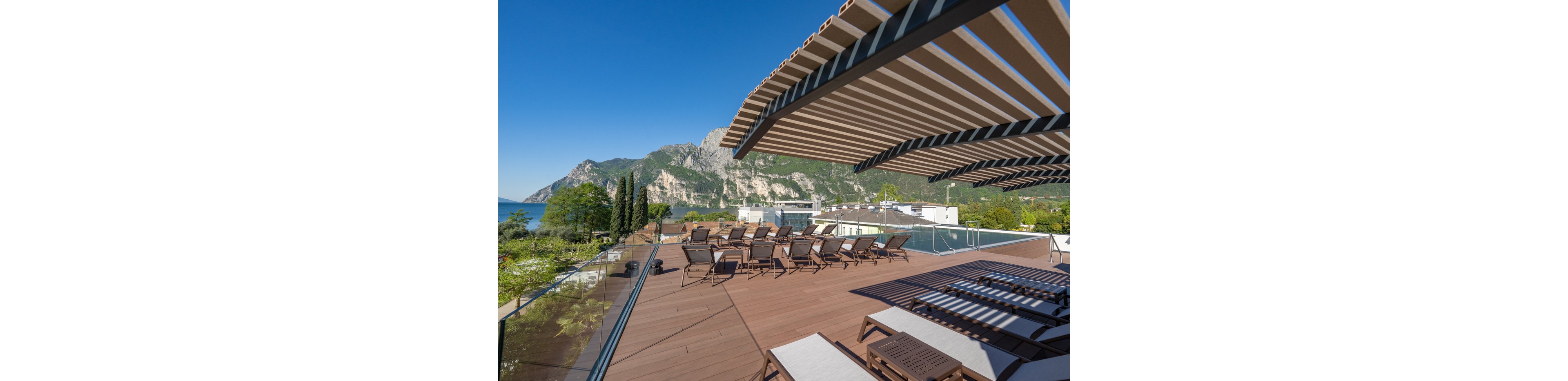 A spacious rooftop terrace features multiple lounge chairs arranged on a wooden deck. A modern slatted pergola casts striped shadows across the seating area. Glass railings frame the terrace, opening onto a scenic view of a lake and dramatic mountains in the background. The clear blue sky and lush greenery create a bright, relaxing vacation atmosphere.