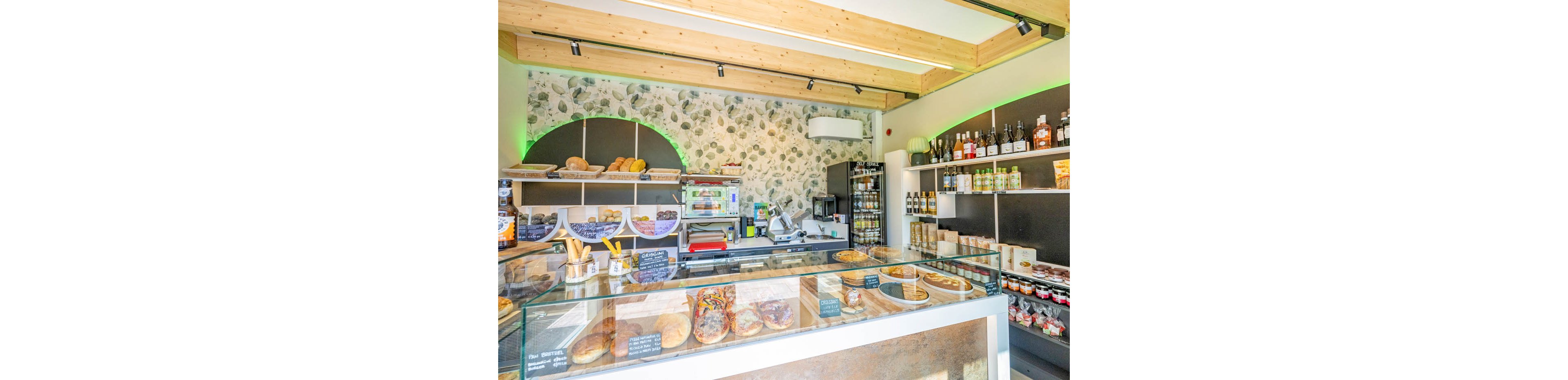 Bright interior of a small artisan bakery with a glass display counter filled with pastries, bread, and savory items. Shelves behind the counter hold loaves of bread, bottled drinks, and various packaged foods. A patterned stone-effect wall and wooden ceiling beams create a warm, contemporary atmosphere. Small chalkboard signs label items in the case, enhancing the cozy, local shop feel.