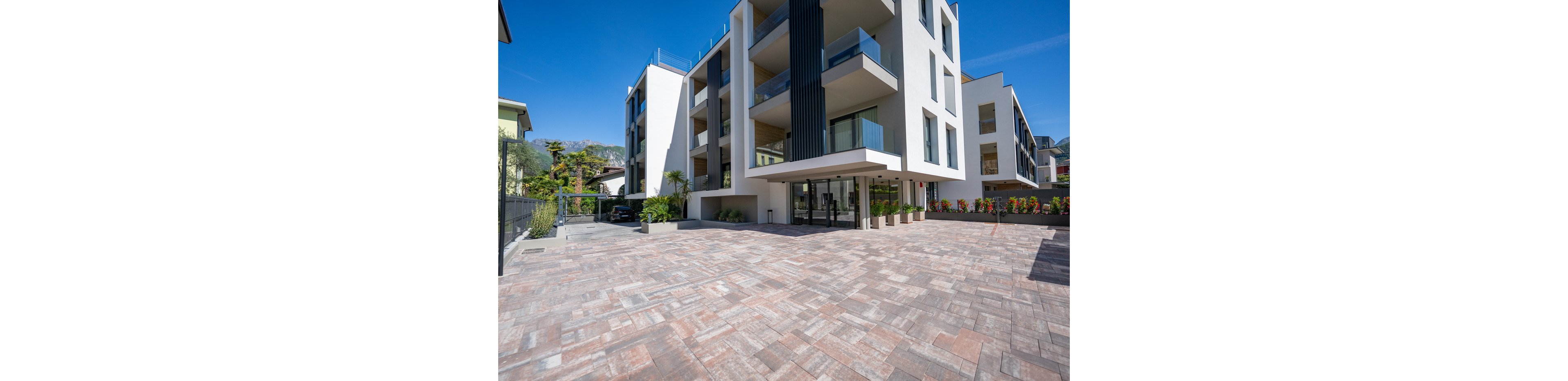 Contemporary multi-story apartment building with large balconies overlooks a wide paved courtyard. The foreground features patterned stone or tile paving leading toward the glass-fronted entrance. Planters with greenery and flowers soften the modern architecture and add color along the perimeter. Clear blue sky and distant hills suggest a sunny, upscale residential neighborhood.