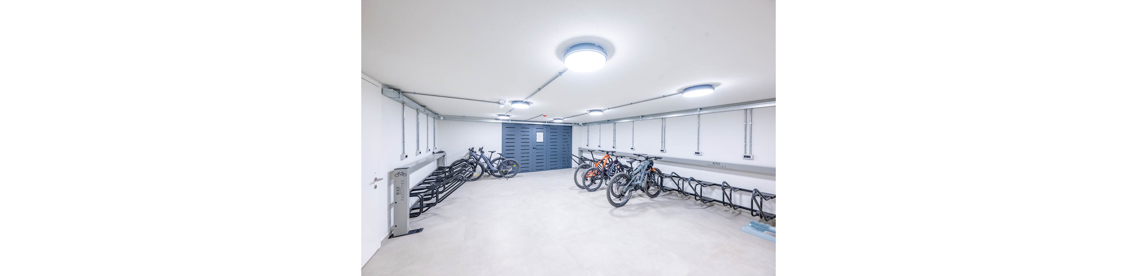 A clean, modern indoor bicycle storage room features several bikes parked in metal racks. The space has white walls, a light floor, and bright circular ceiling lights providing even illumination. A large gray storage or utility cabinet stands at the far end of the room. The overall scene suggests secure, organized parking for residents or office workers.