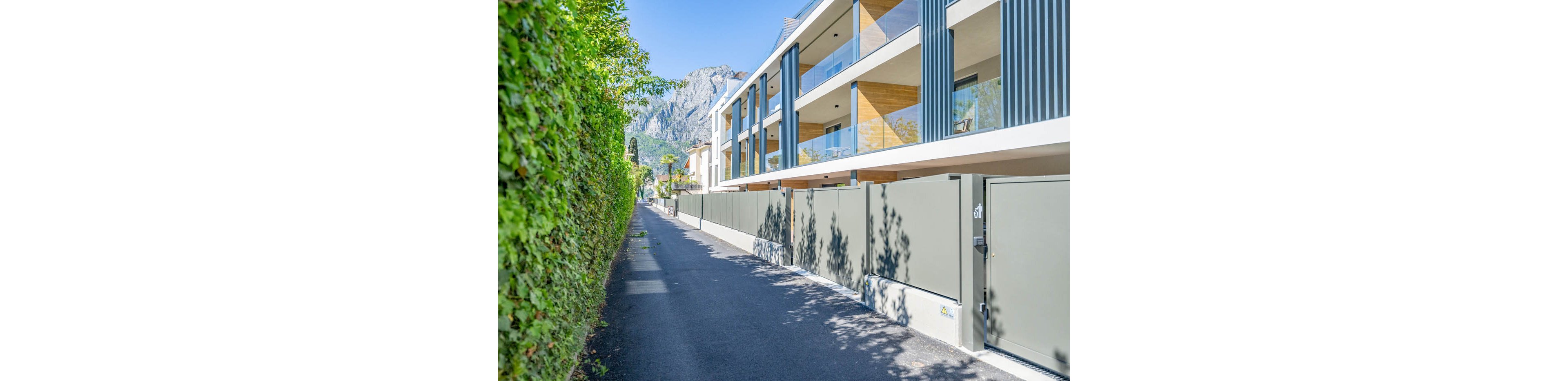 A contemporary multi-story apartment building lines a narrow paved pathway on a bright sunny day. The structure features glass balconies, wood accents, and vertical metal panels facing the street. A tall hedge borders the opposite side of the path, casting soft shadows on the pavement. Mountains rise in the background under a clear blue sky, adding a scenic natural backdrop to the residential scene.