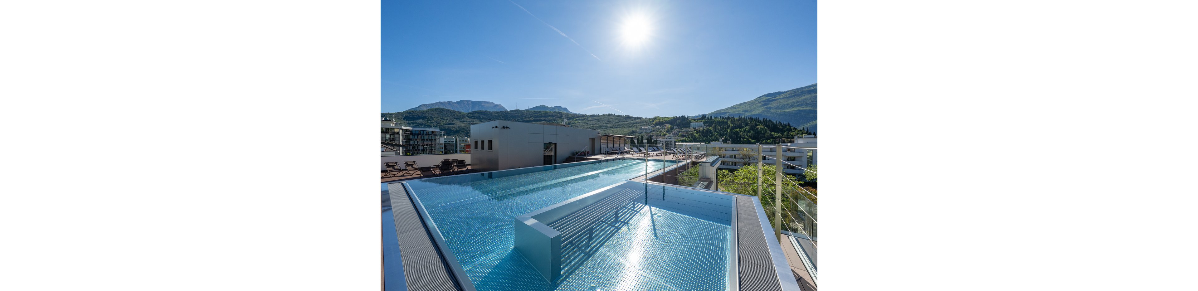 A sleek rooftop infinity pool reflects the bright midday sun. The pool sits atop a contemporary building with lounge chairs and railings along the deck. In the background, green hills and distant mountains frame the urban setting. The clear blue water and cloudless sky create a calm, luxurious atmosphere.
