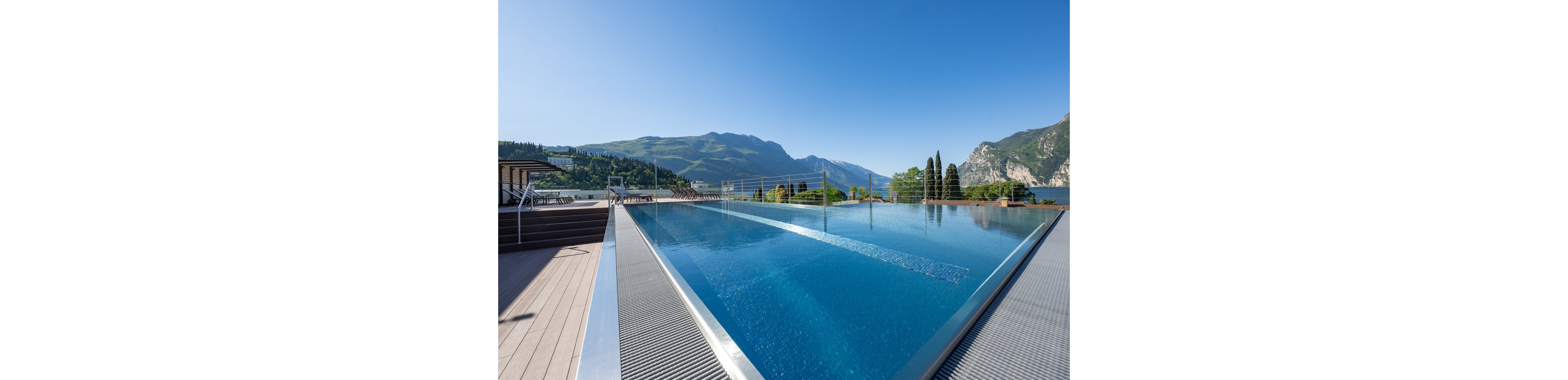 A modern rooftop infinity pool stretches toward a scenic mountain landscape. Clear blue water reflects the bright sky and surrounding peaks. Wooden decking and metal railings frame the pool area, suggesting a contemporary resort or hotel setting. In the distance, a calm lake and lush green hills complete the tranquil outdoor scene.