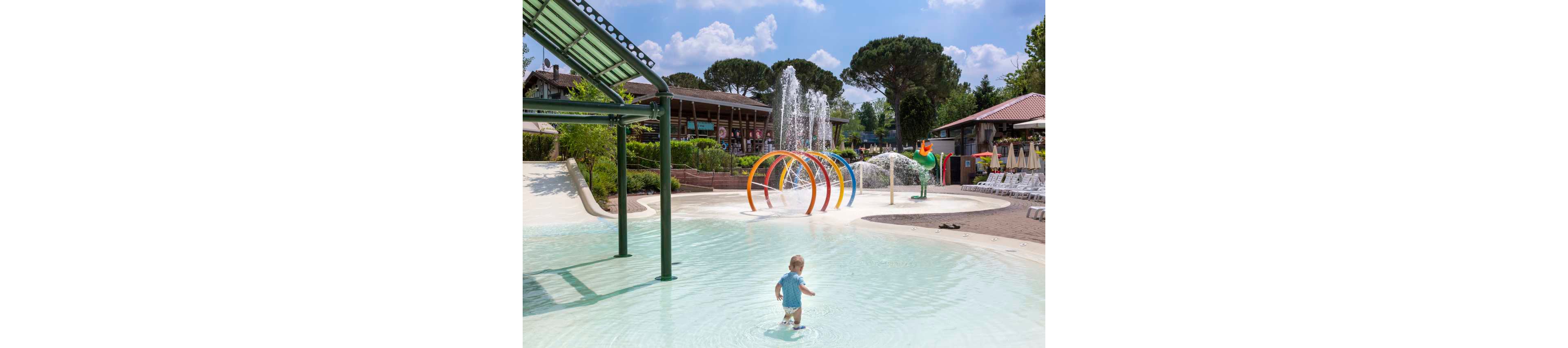 A small child walks through a shallow splash pool at an outdoor water park. Colorful circular spray arches and tall water fountains create a playful backdrop. Surrounding the pool are lounge chairs, umbrellas, and landscaped greenery under a bright blue sky. The scene feels relaxed and family-friendly, suggesting a warm summer day at a resort or campground.