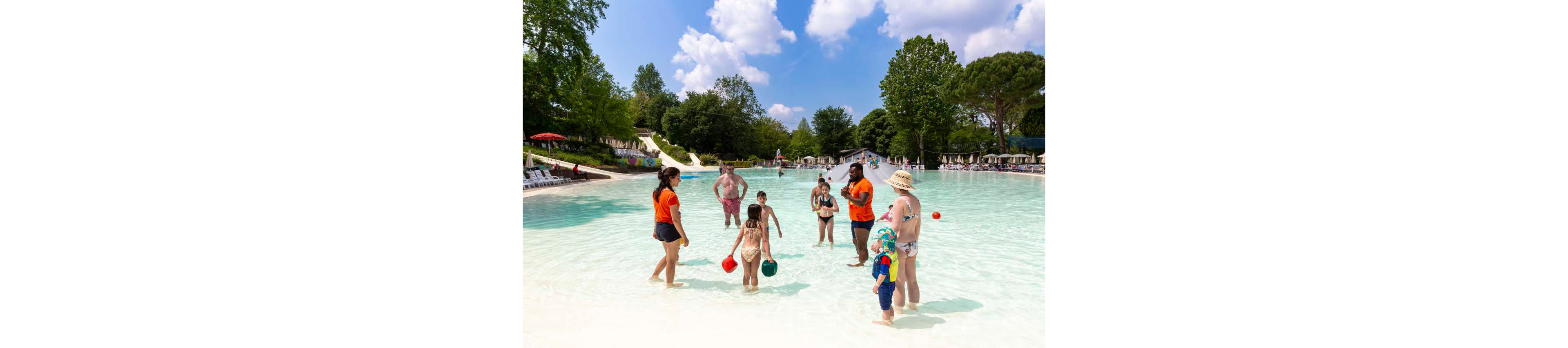 Several families enjoy a sunny day in a shallow outdoor swimming pool. Children play with colorful buckets and balls while adults stand nearby in the water supervising. Two staff members in matching orange shirts interact with the kids, suggesting an organized activity or resort setting. Lush green trees, lounge chairs, and water slides are visible in the background under a bright blue sky with scattered clouds.