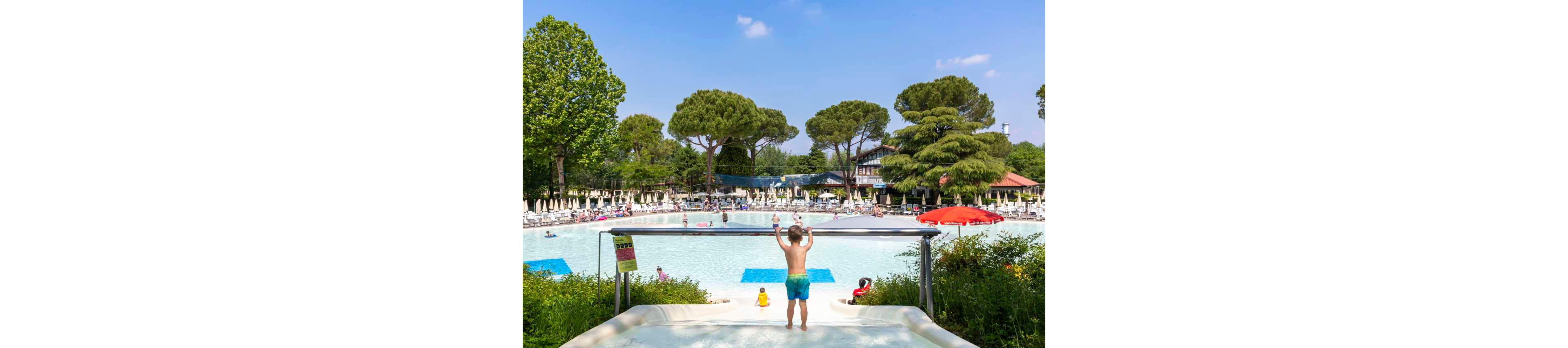 A young child stands at the top of a waterslide overlooking a large outdoor pool. The scene is set in a busy water park or resort with many people relaxing on loungers around the pool. Tall green trees and landscaped gardens surround the area, creating a lush summer atmosphere. Bright sunlight and clear blue sky enhance the cheerful, family-friendly vacation setting.