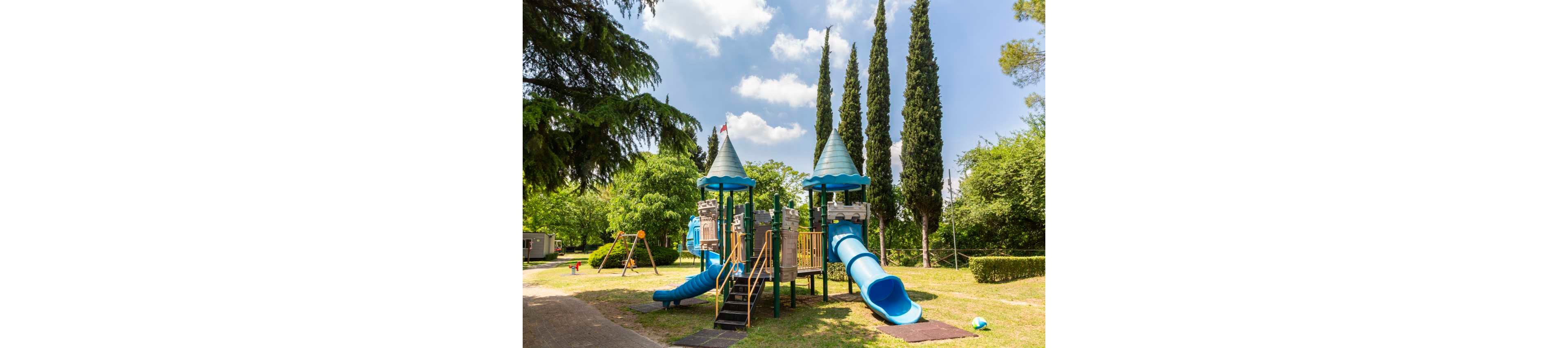 A colorful playground structure with blue slides and towers stands in a green park. The play area includes climbing platforms, stairs, and a tunnel slide surrounded by grass and trees. In the background, there is a swing set and additional park equipment under a bright blue sky with scattered clouds. Tall cypress trees and other lush vegetation frame the scene, creating a peaceful outdoor setting.