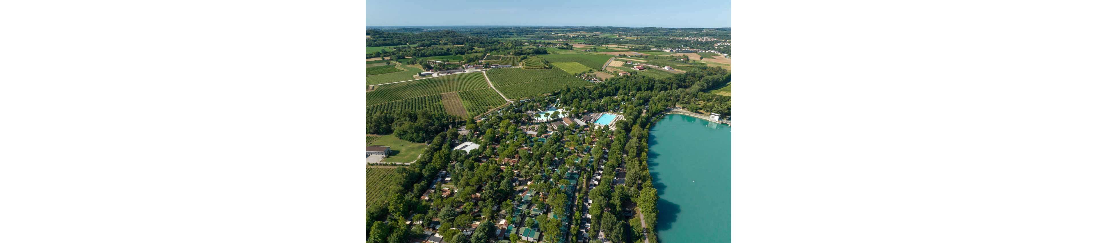 A wide aerial view shows a large campground nestled among dense trees beside a turquoise lake. Numerous cabins, tents, and RVs are arranged along narrow roads, with swimming pools and recreation areas visible near the center. Surrounding the campsite are expansive green fields and farmland stretching to the horizon. The scene suggests a peaceful rural holiday destination with abundant nature and outdoor activities.