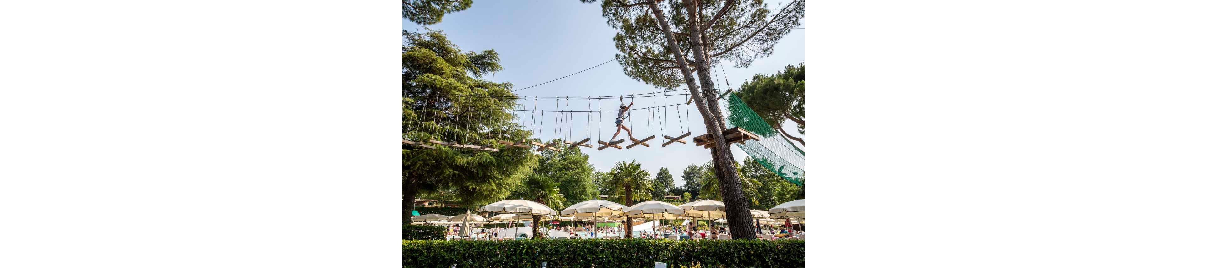 A child wearing a helmet and harness crosses a high ropes course suspended between trees. The adventure course is set above a busy outdoor pool area filled with sun umbrellas and people relaxing. Lush green trees and foliage surround the scene, creating a vibrant summer atmosphere. The image conveys leisure, adventure, and family vacation fun in a resort-like setting.