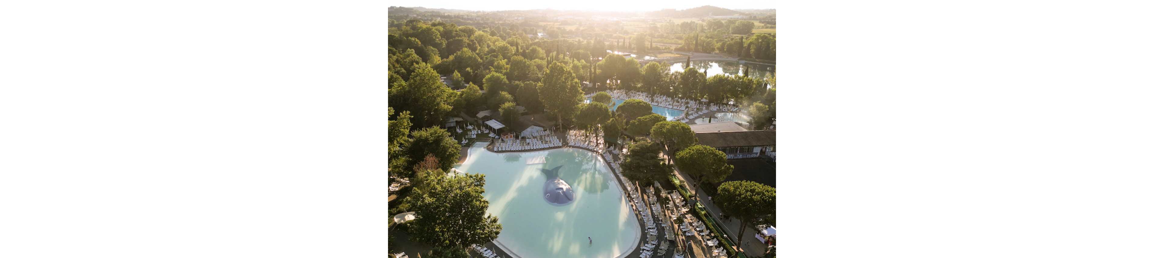 A large resort-style lagoon pool is surrounded by sun loungers and tall trees in a lush green landscape. The aerial view shows multiple swimming pools and a central shallow pool with a whale-shaped play structure. Soft golden sunlight filters through the trees, creating long shadows and a tranquil atmosphere. In the distance, rolling hills and a river or lake complete the scenic vacation setting.