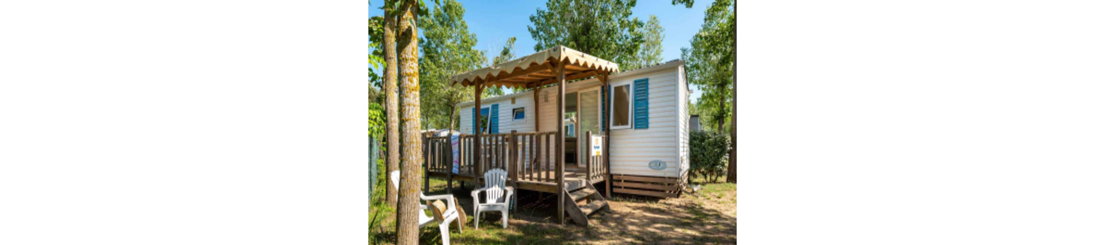 A compact mobile home with blue shutters stands in a sunny, wooded campsite. The structure features a raised wooden deck with a canopy providing shade over the entrance. White plastic chairs are arranged on the ground and on the terrace, suggesting a relaxed outdoor seating area. Tall trees and green foliage surround the home, creating a peaceful vacation atmosphere.