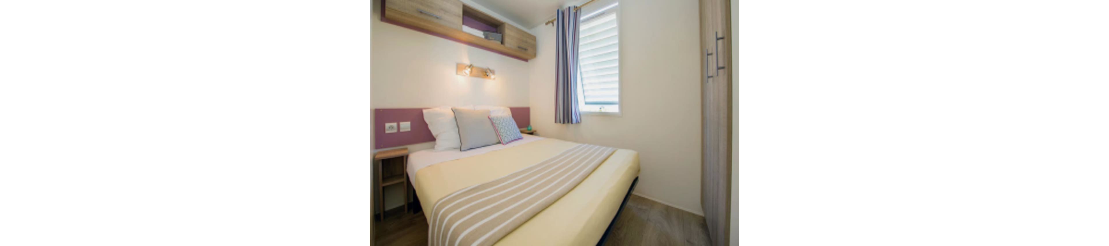 A compact modern bedroom features a neatly made double bed with striped beige and white bedding. Soft pillows in white, gray, and patterned fabric add a touch of color against a light wall with a purple accent panel. Built-in wooden cabinets and a small bedside table provide storage in the narrow space. A window with striped curtains and a louvered blind lets in natural light, creating a bright and inviting atmosphere.