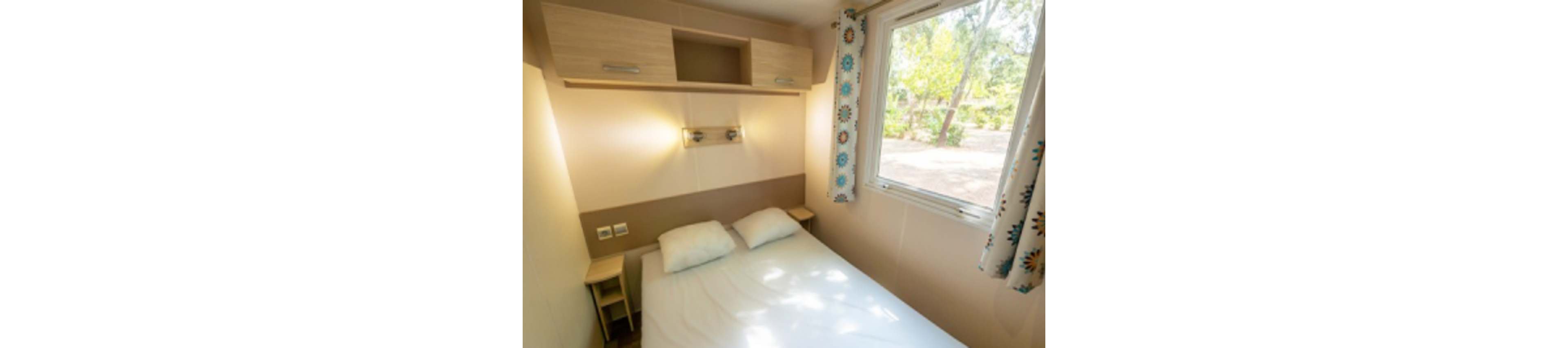 A small, neatly made bed sits in a compact bedroom with light wood furniture. Above the bed, wall-mounted cabinets and twin reading lights provide storage and illumination. A large window with patterned curtains lets in natural light and offers a view of trees outside. The room has a clean, minimalist design suitable for a vacation cabin or mobile home.