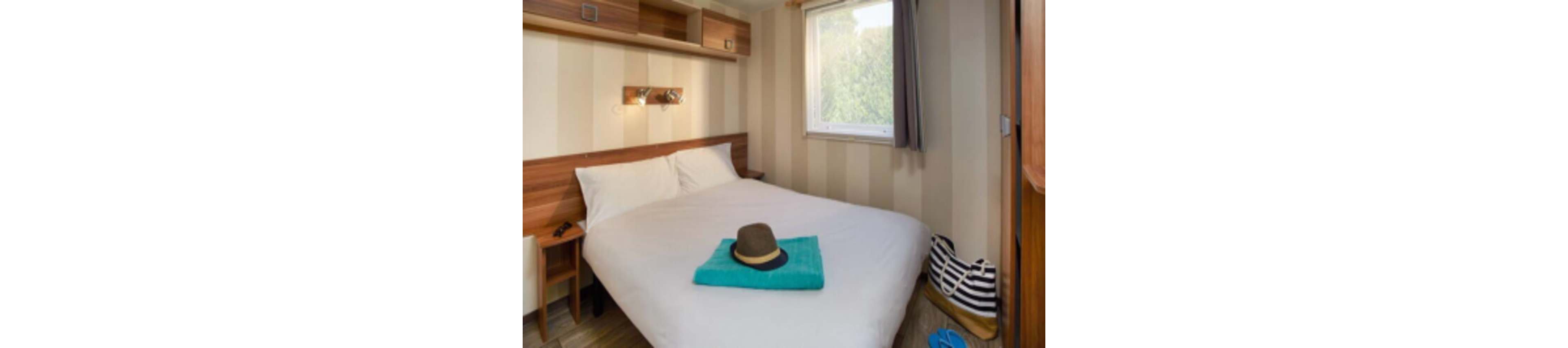 A small, cozy bedroom features a neatly made double bed with white linens. A folded turquoise towel and a brown hat are placed at the foot of the bed, suggesting a vacation or weekend getaway. Built-in wooden shelves and wall lights frame the head of the bed, while a window lets in natural light and greenery from outside. A striped bag and blue sandals sit on the bench by the bed, adding to the relaxed travel atmosphere.