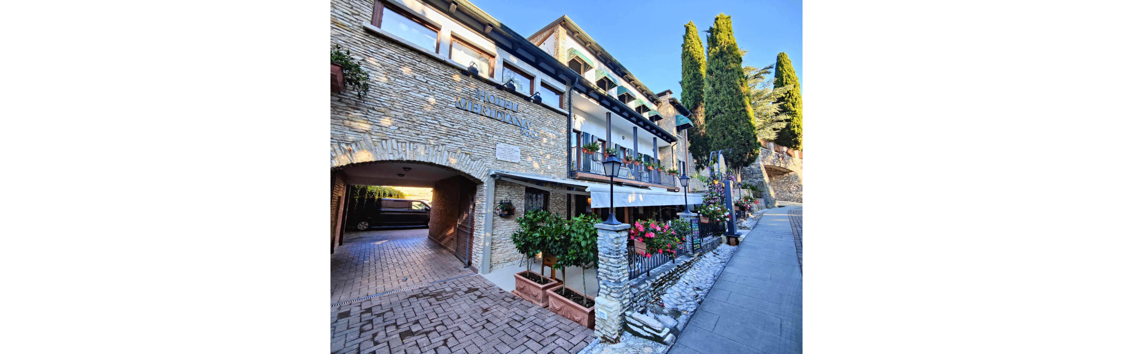 A quaint stone hotel with balconies and flower boxes lines a sloped hillside street. The building features arched entrances, white railings, and colorful potted plants along the facade. Tall evergreen trees rise behind the structure, adding to the scenic village atmosphere. Warm sunlight illuminates the cobblestone driveway and walkway, creating a welcoming, tranquil mood.
