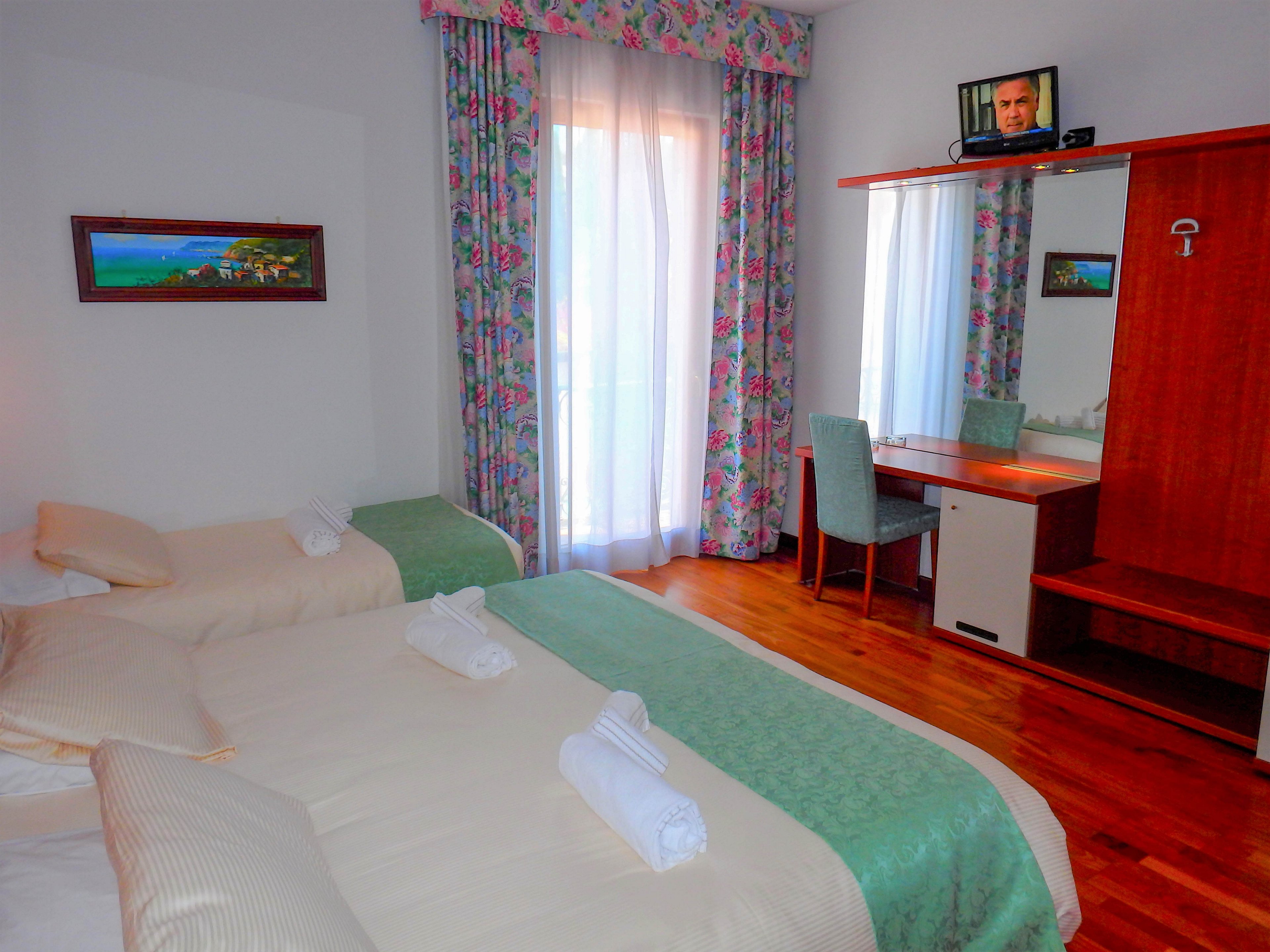 A cozy hotel room features a double bed and a single bed neatly made with light bedding and rolled white towels. Natural light streams through sheer curtains covering a tall window, framed by floral drapes. A wooden desk with a mirror, chair, and small television sits against one wall, adding functionality to the space. Warm wooden flooring and a colorful wall painting contribute to the inviting, homey atmosphere.