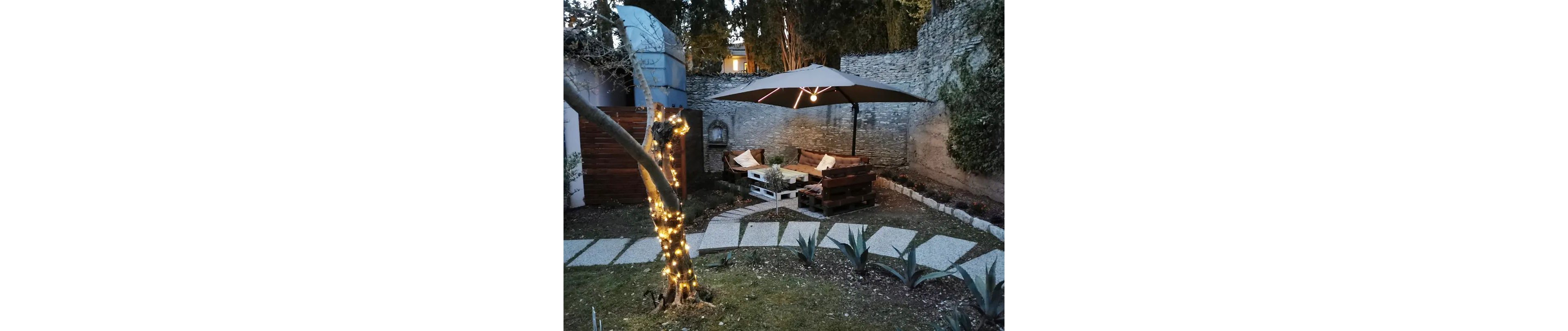 An inviting backyard seating area features wooden pallet sofas arranged around a low table beneath a large patio umbrella. The space is set against a stone wall with trees and shrubs providing a natural backdrop. A small tree wrapped in warm string lights adds a soft glow to the garden at dusk. Stepping stones lead through the yard past agave-like plants, enhancing the intimate outdoor lounge atmosphere.