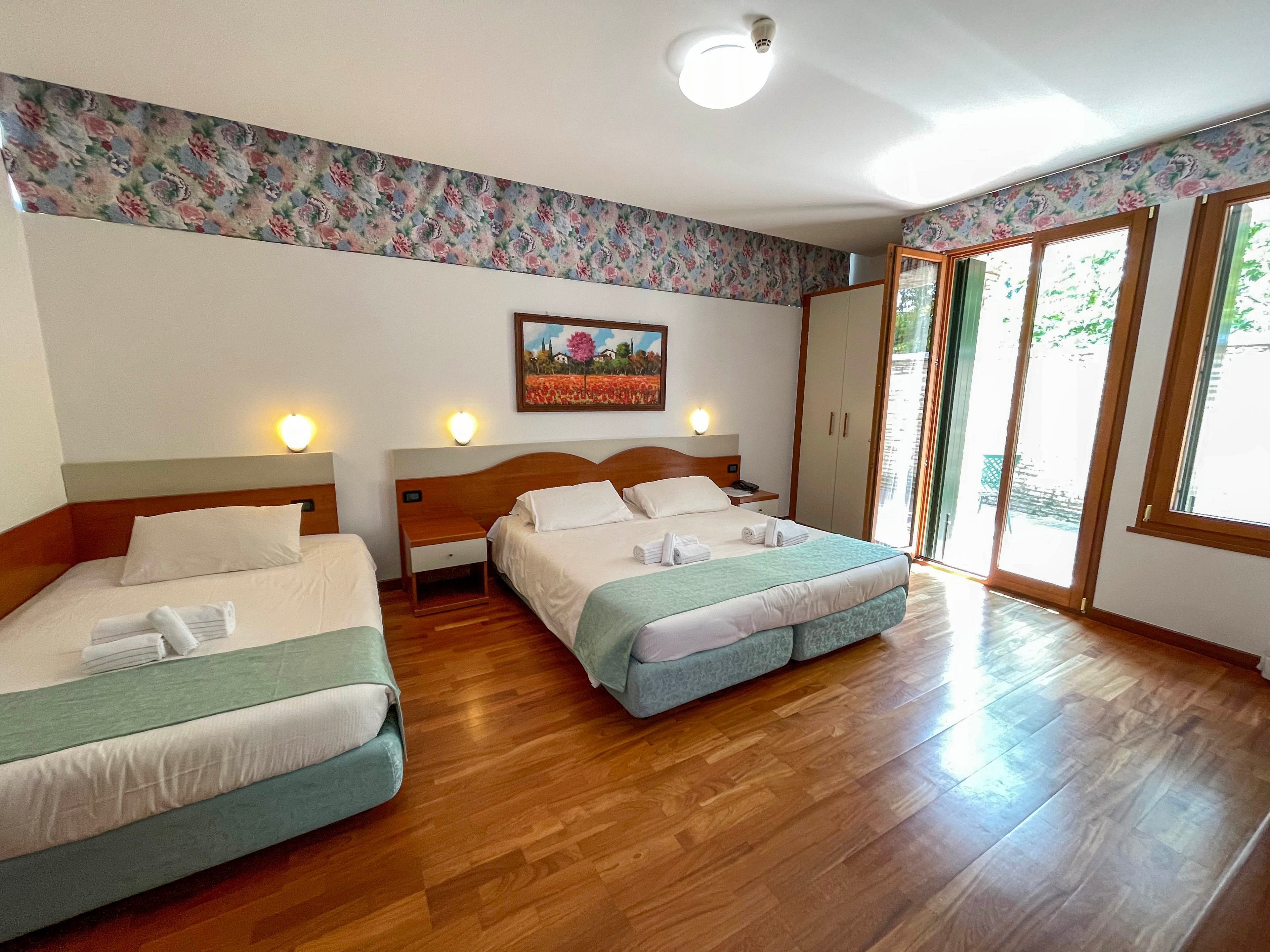 A spacious hotel room features three neatly made beds with white linens and light green throws. The room has wooden floors and a large windowed door that opens to an outdoor area, allowing natural light to fill the space. Soft wall-mounted lights and a colorful floral painting above the central bed add warmth and character. Folded towels are placed on each bed, suggesting readiness for guests and a clean, welcoming atmosphere.