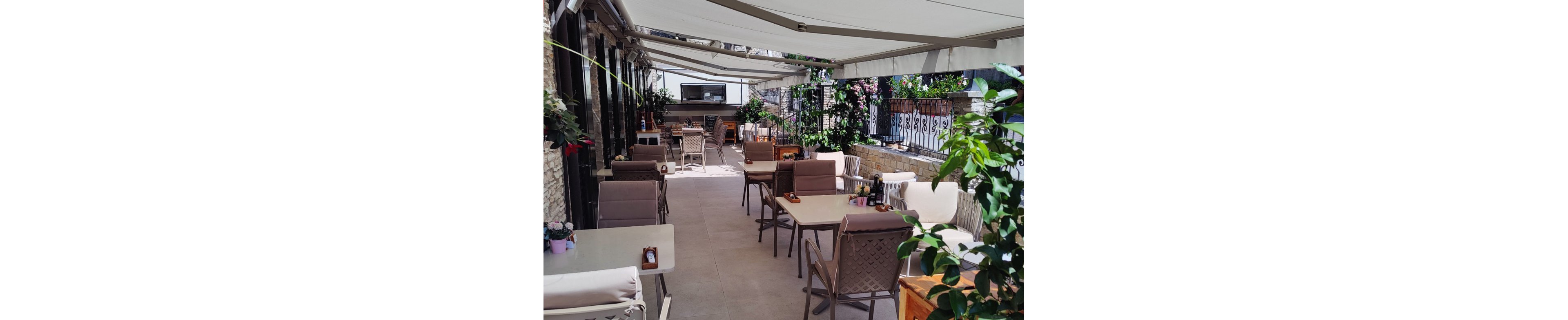 An empty outdoor café terrace is arranged with neatly spaced tables and chairs under large white awnings. The setting appears bright and sunny, with soft shadows cast on the light stone floor. Potted plants and greenery line the sides, adding a relaxed, natural atmosphere. In the background, a bar or service counter is visible, enhancing the impression of a quiet, modern dining space.