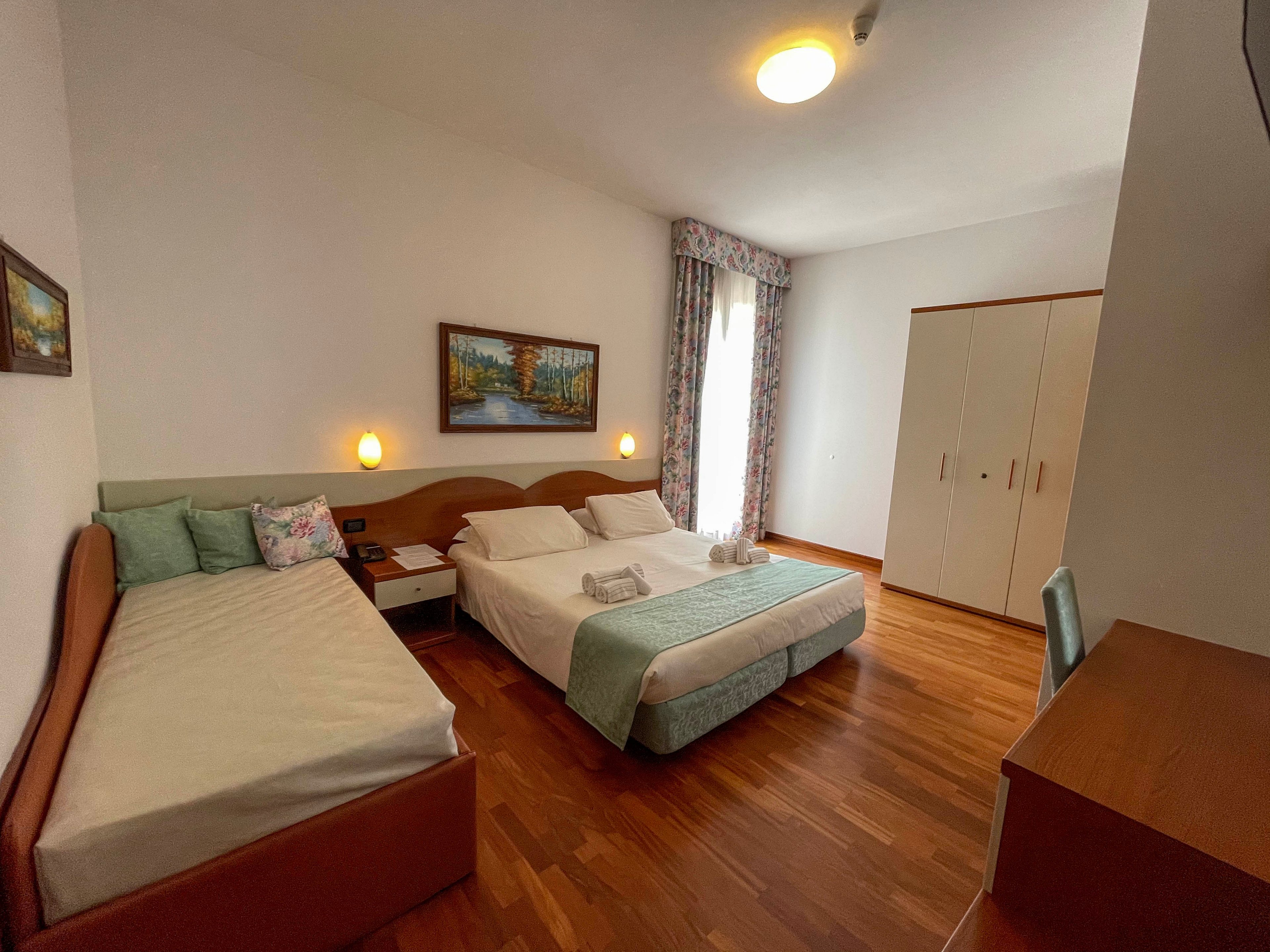 A bright hotel room features a neatly made double bed and a single bed with folded towels placed on top. The space includes wooden flooring, a white wardrobe, and a desk with a chair. Soft lighting from wall sconces and a ceiling light creates a warm atmosphere. A large window with patterned curtains and framed artwork on the wall add decorative touches to the simple, cozy interior.