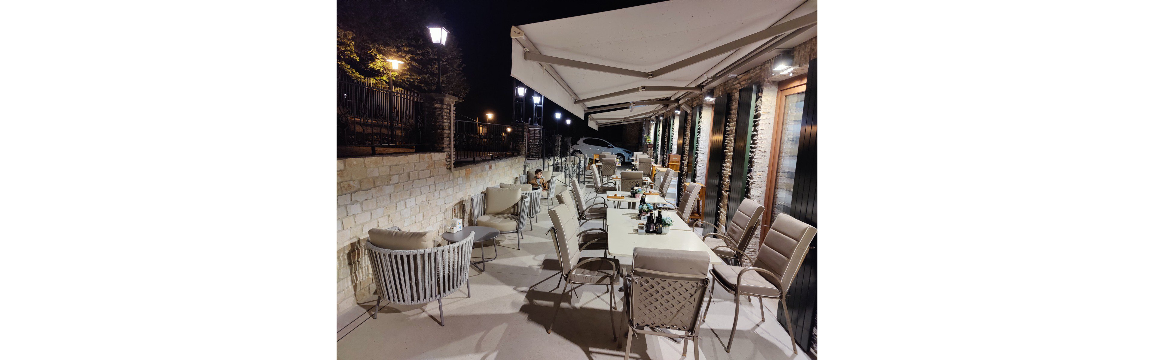 An outdoor terrace dining area is set up at night with a long table surrounded by cushioned chairs. The space is covered by large retractable awnings attached to a stone and brick building. Warm lights from wall fixtures and street lamps illuminate the patio and nearby parked car. A few bottles and glasses remain on the table, suggesting a recently finished gathering. The overall atmosphere feels relaxed and inviting for evening socializing.