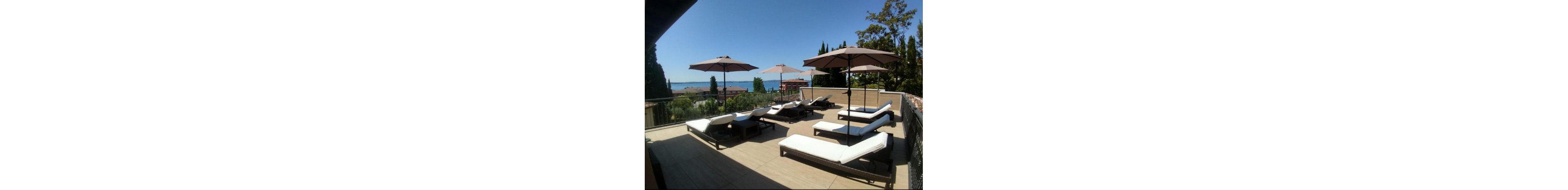 A spacious rooftop terrace features several white cushioned lounge chairs arranged under large beige umbrellas. The area is enclosed by a metal railing and overlooks a scenic town with red-roofed buildings. In the distance, a calm blue body of water and a clear sky create a relaxing vacation atmosphere. Tall trees frame the view, adding greenery to the bright, sunlit setting.