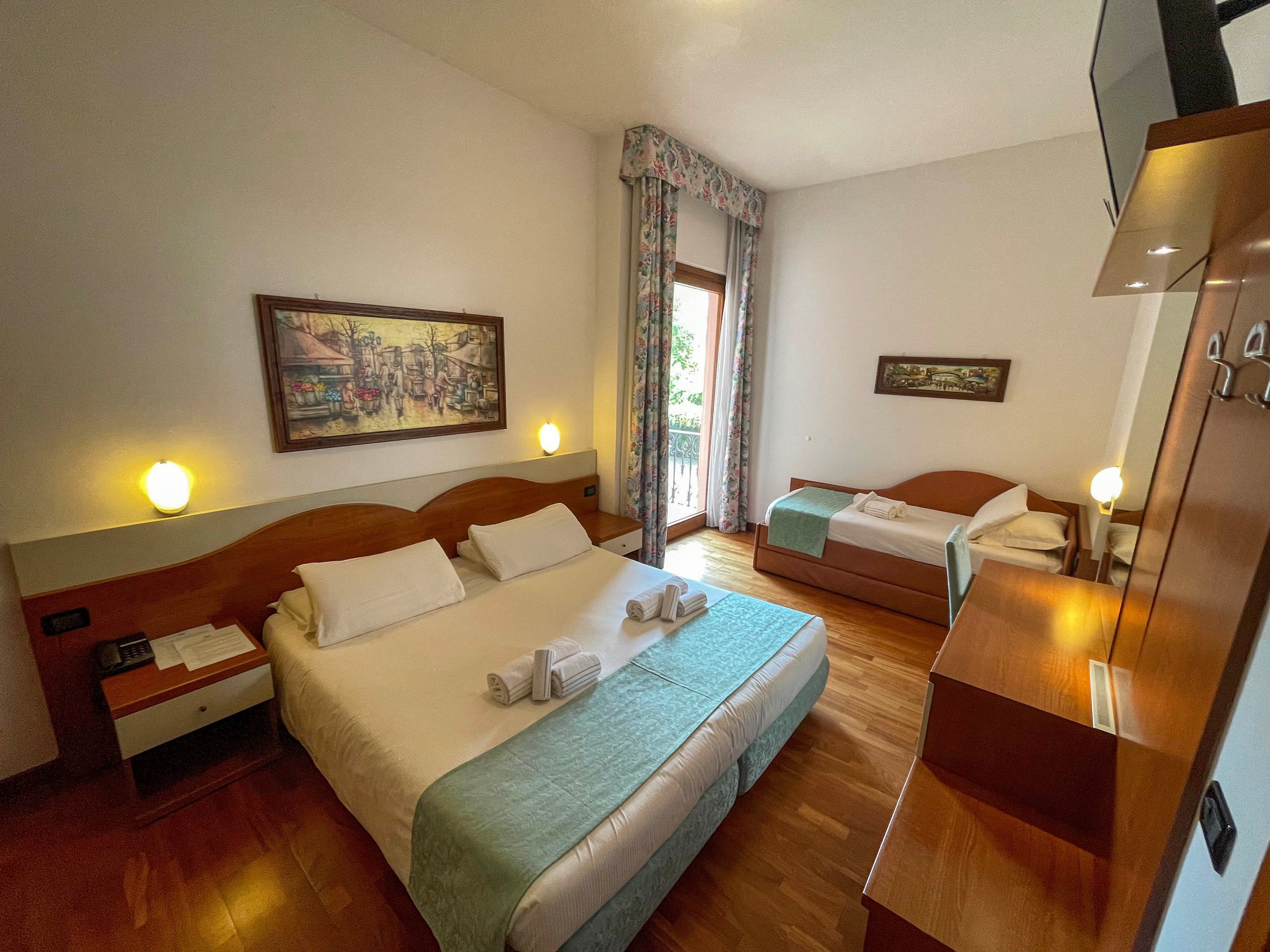 A neatly arranged hotel room features a large double bed in the foreground and a single bed along the side wall. The space has wooden floors, light-colored walls, and coordinated bedding in white and soft turquoise tones. Wall-mounted lamps, framed artwork, and a balcony door with patterned curtains add warmth and character. Towels are folded on the beds, and built-in wooden furniture and a wall-mounted television complete the cozy, modern accommodation.