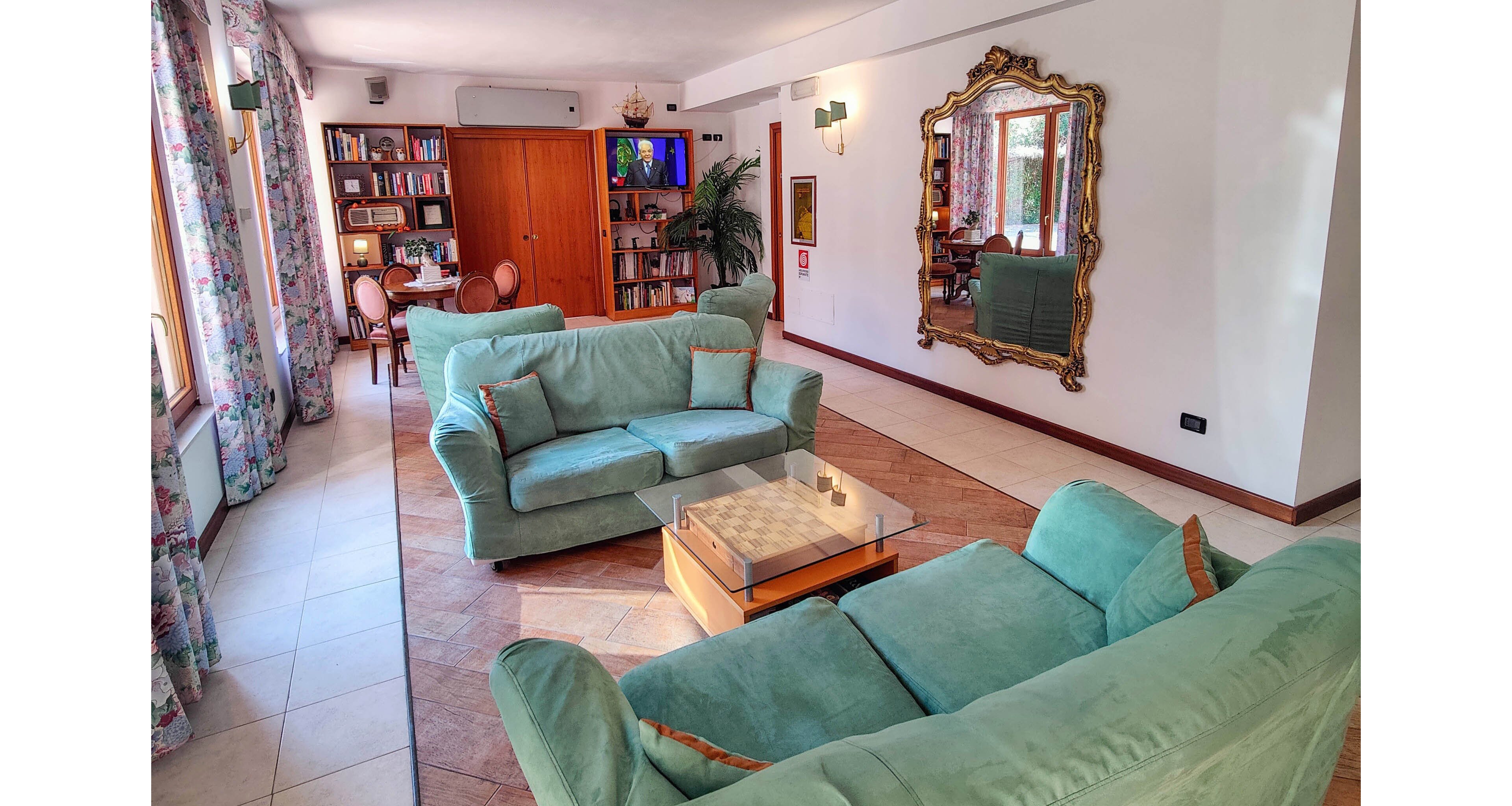 A spacious living room features two teal sofas arranged around a wooden coffee table. Large windows with patterned curtains allow natural light to fill the space. In the background, a wall of bookshelves, a television, and wooden cabinetry create a cozy reading and entertainment area. A large ornate gold-framed mirror and indoor plants add decorative touches to the white walls and tiled floor.