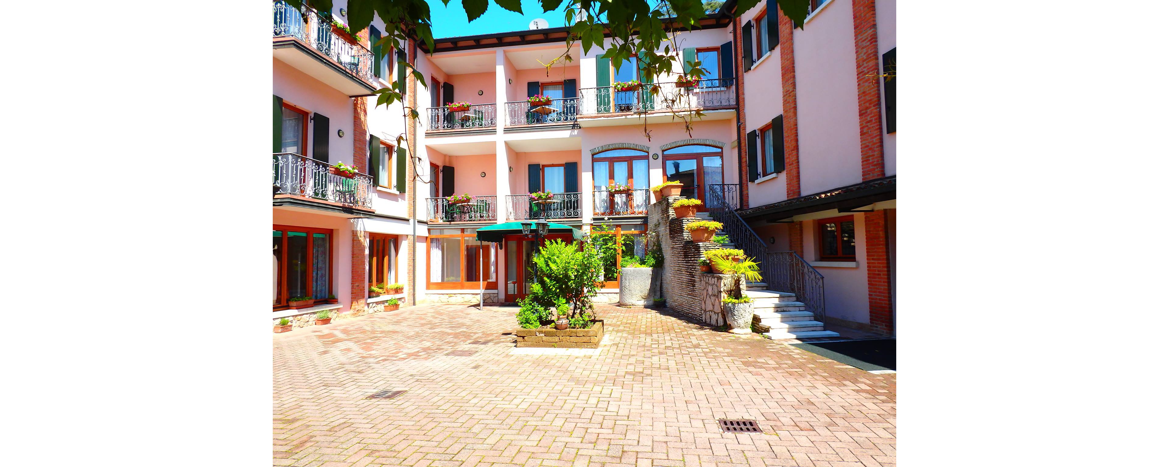 A bright residential courtyard is surrounded by multi-story buildings with pastel facades and wrought-iron balconies. Large windows and doors open onto the courtyard, creating an inviting communal space. A curved stone staircase with potted plants leads to the upper level, adding architectural interest. The sunlit brick paving and greenery give the scene a warm, Mediterranean atmosphere.