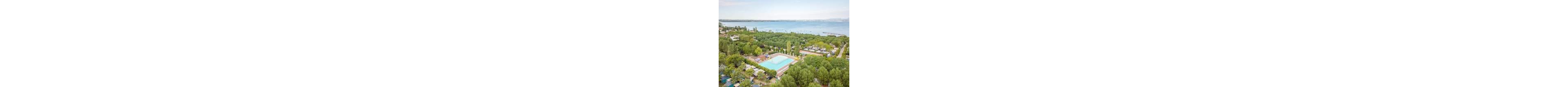 A large outdoor swimming pool sits at the center of a lush, tree-filled campsite near a wide lake. The aerial view reveals rows of camping plots, caravans, and small buildings surrounded by greenery. In the background, the calm lake stretches toward distant hills under a light, hazy sky. The overall scene suggests a peaceful holiday destination focused on nature and outdoor recreation.