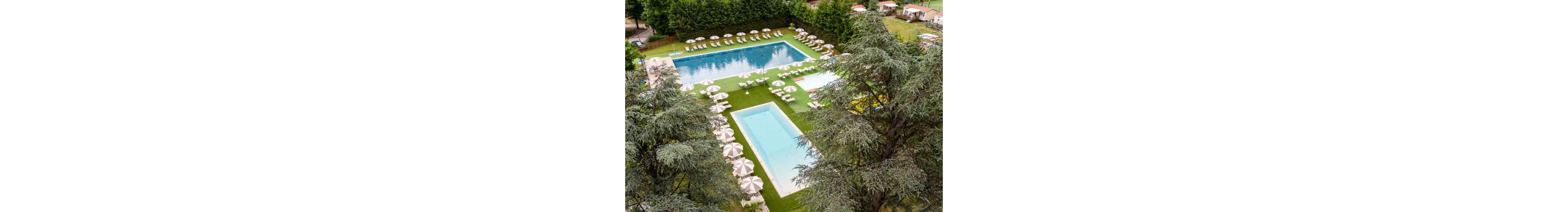 An aerial view shows a landscaped outdoor swimming pool complex surrounded by tall trees and greenery. Multiple rectangular pools of different sizes are arranged on a manicured lawn. Numerous sun loungers and umbrellas are neatly lined up around the pools, suggesting a resort or holiday park setting. Small buildings and cabins are visible in the background, adding to the vacation atmosphere.