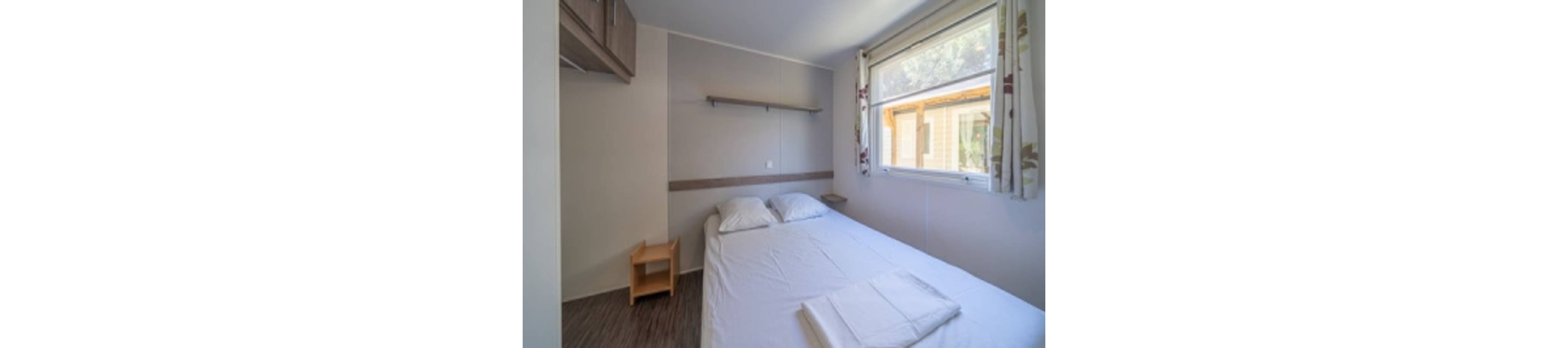 A small, minimalist bedroom features a neatly made double bed with white linens and two pillows. Natural light enters through a large window with patterned curtains, offering a view of another building outside. A simple wooden nightstand and wall shelf add functional storage to the compact space. The overall atmosphere is clean, bright, and uncluttered, suitable for a vacation rental or guest room.