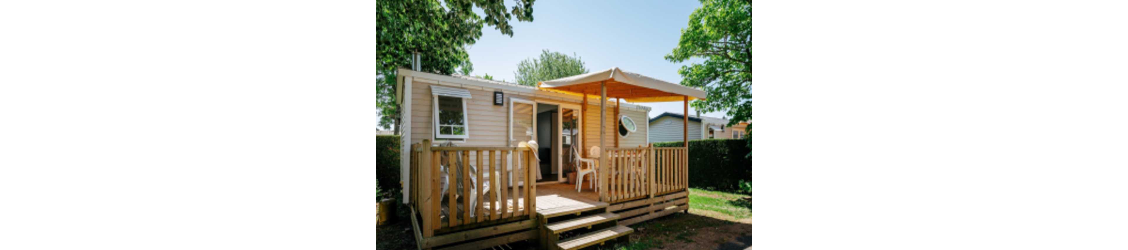 A compact mobile home with light-colored siding features a small wooden deck and steps leading to the entrance. A pergola-style canopy shades the outdoor seating area, where white plastic chairs and a table are arranged. The home is set in a green, tree-filled environment with neighboring units visible in the background. Bright daylight and lush foliage create a relaxed, vacation-like atmosphere.