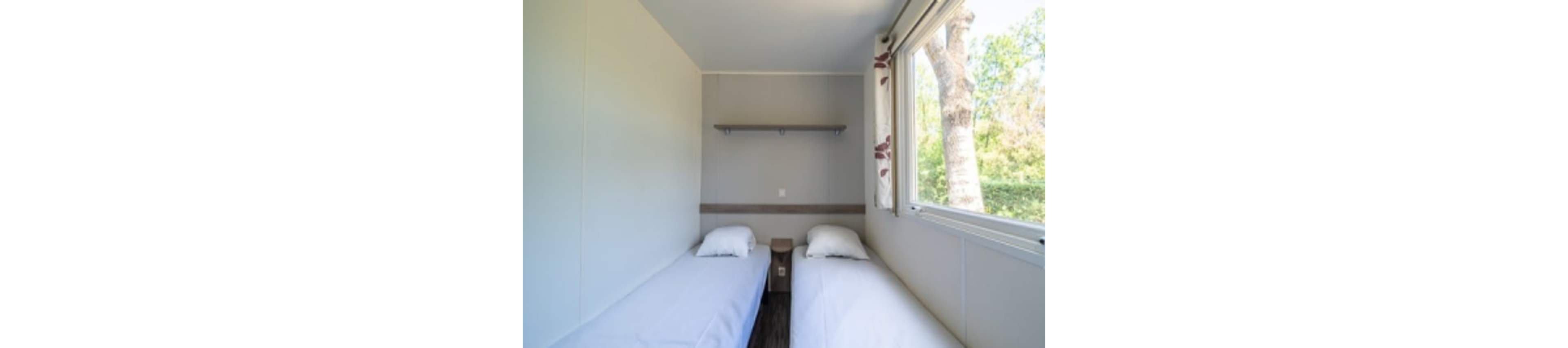 A small bedroom features two single beds with white linens separated by a narrow bedside table. The room has plain light-colored walls and a simple floating shelf above the headboards. A large window on the right lets in natural light and offers a view of trees and greenery outside. The overall atmosphere is clean, minimal, and functional, suitable for a guest room or vacation rental.