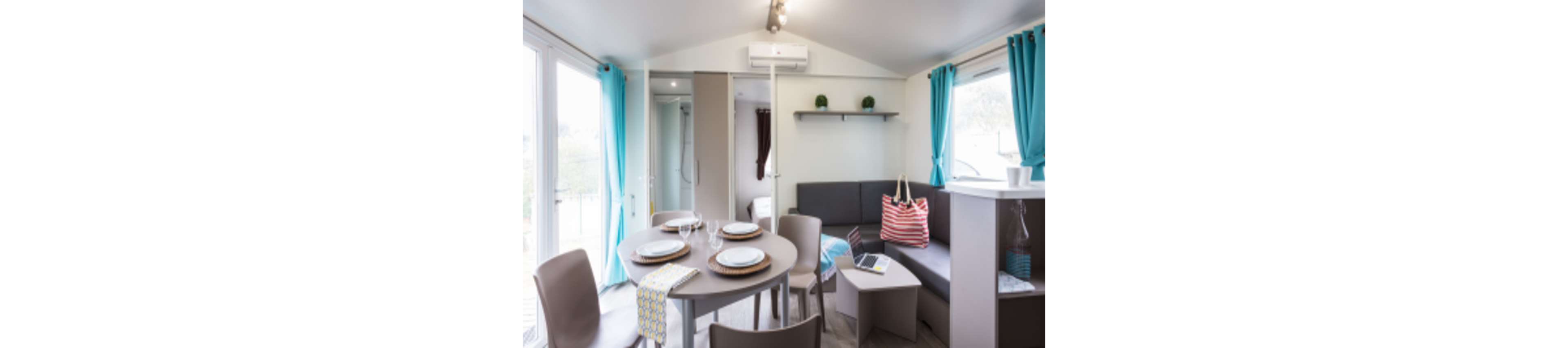 Compact living and dining area inside a modern mobile home features a round table set with plates and glasses. A gray corner sofa with a striped beach bag and open laptop sits beside a small coffee table. Large windows with turquoise curtains bring in natural light, and a doorway reveals a bedroom and bathroom area. The space feels clean, contemporary, and ready for a holiday stay.